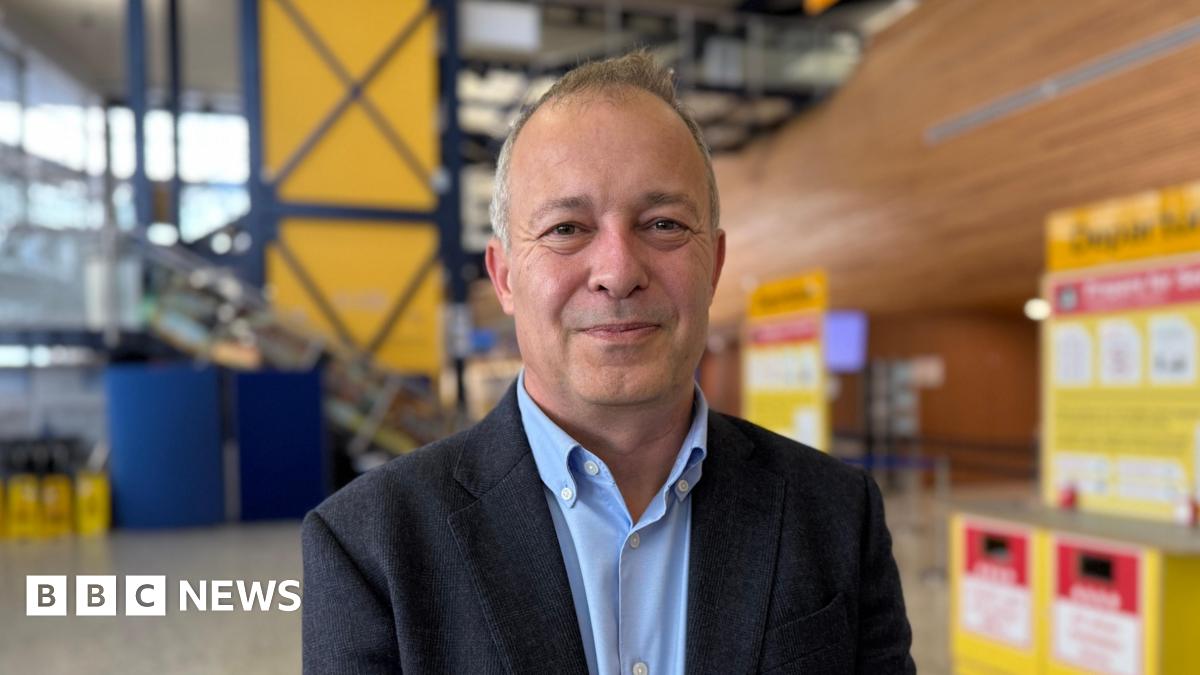 Nico Bezuidenhout, a man with short grey hair, wearing a light blue shirt and a dark blazer. He is smiling directly at the camera, while standing in the departures hall at Guernsey Airport.