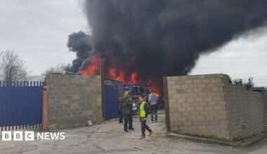 A thick black cloud of smoke above a fire burning behind a breeze block wall. The fire looks to be on top of a scrap heap of cars, with a blue burnt out car on top.