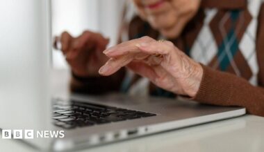 Stock image of a woman with short hair wearing a grey/green dressing gown with white spots on it, looking out towards a window covered in vertical blinds