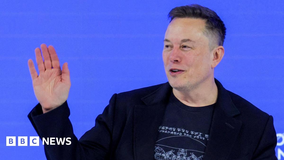 Elon Musk waving to a crowd from a stage wearing a black blazer and a black t-shirt.