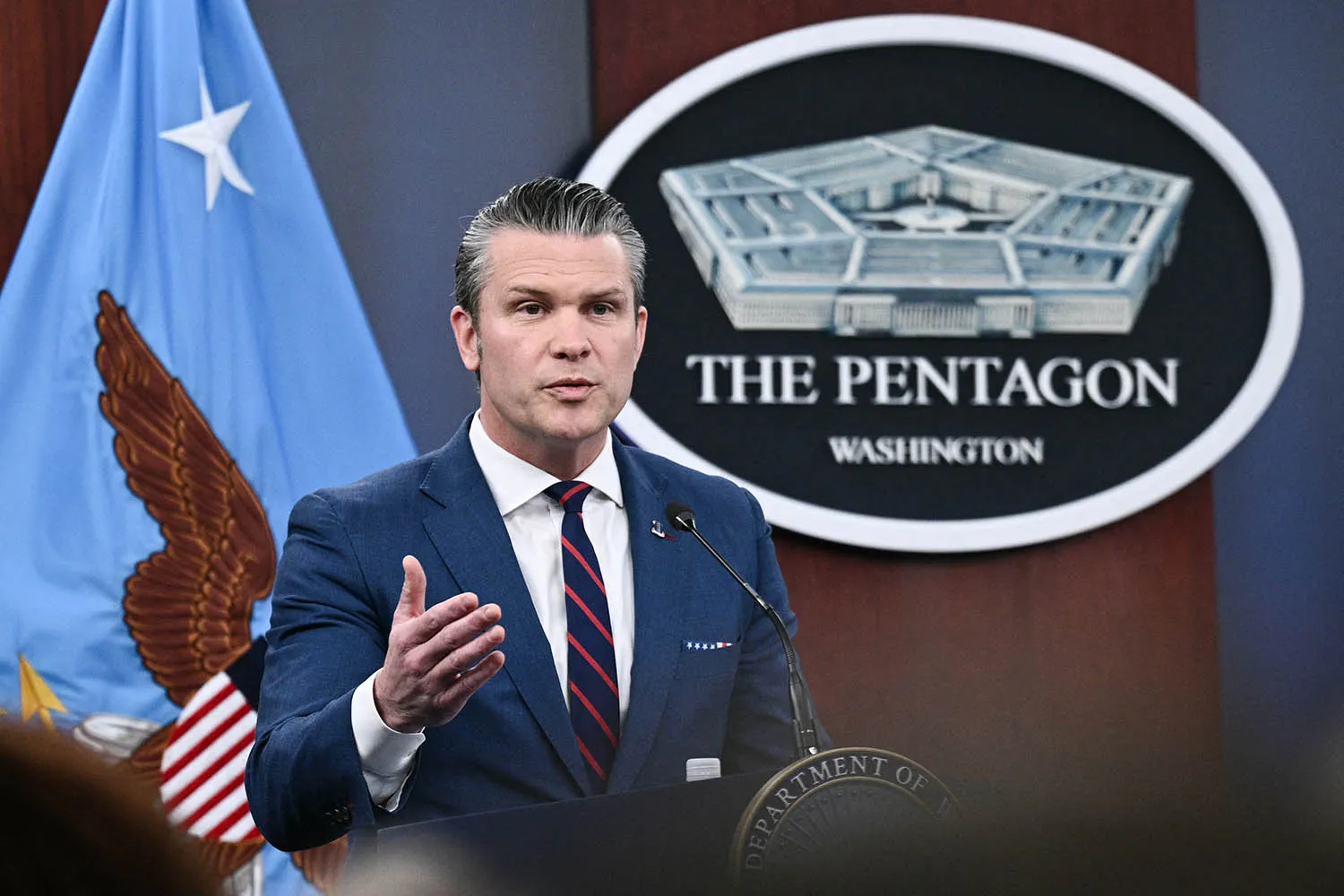 Hegseth, a man in a blue suit and striped tie, speaks at a podium. Behind him is a large wall plaque that reads "THE PENTAGON WASHINGTON" featuring an illustration of the building, flanked by a light blue flag with an eagle.