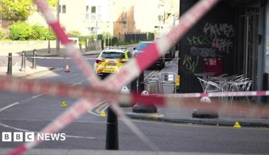 Red police tape crosses the road. Silver chairs are on the pavement opposite, inside the police cordon with a police car with fluorescent yellow marking parked beyond it.