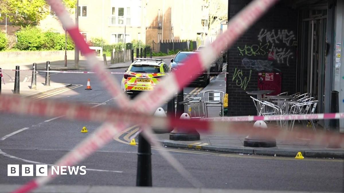 Red police tape crosses the road. Silver chairs are on the pavement opposite, inside the police cordon with a police car with fluorescent yellow marking parked beyond it.