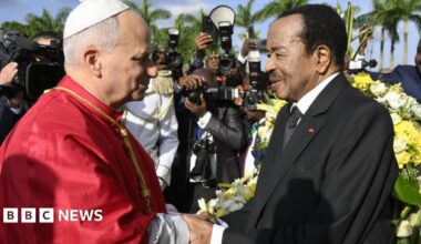 Pope Leo in a red cape shakes the hand of President Paul Biya in a black suit with a white dotted tie. Behind them are banks of photographers and a floral arrangement.
