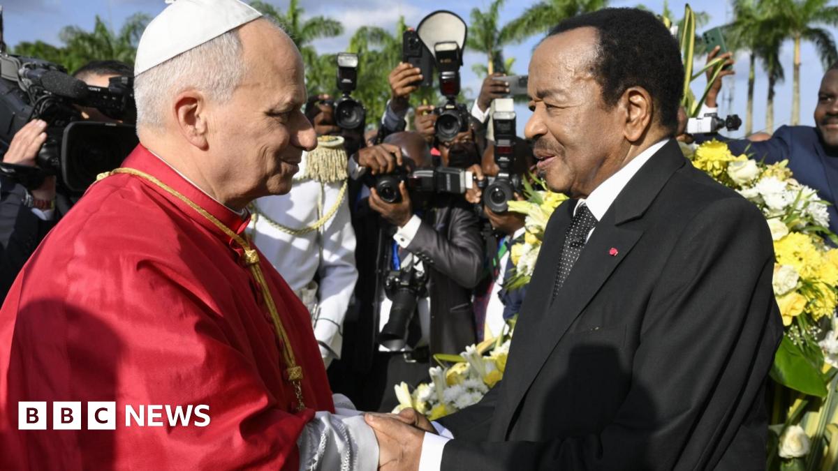 Pope Leo in a red cape shakes the hand of President Paul Biya in a black suit with a white dotted tie. Behind them are banks of photographers and a floral arrangement.
