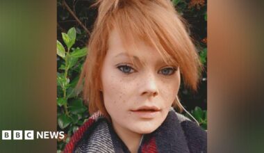 A selfie of a young woman, who has blue almond-shaped eyes, freckles and strawberry blonde hair, which is tied up. She is wearing a scarf and standing in front of greenery.