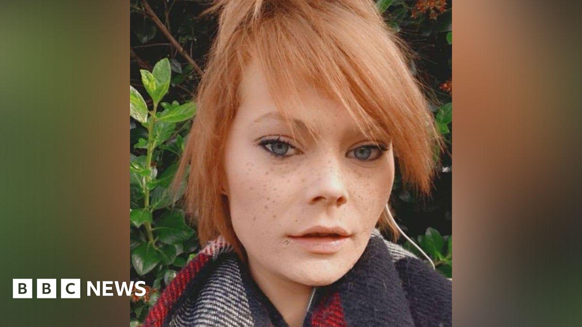 A selfie of a young woman, who has blue almond-shaped eyes, freckles and strawberry blonde hair, which is tied up. She is wearing a scarf and standing in front of greenery.