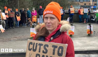 Dr Melissa Ryan wearing an orange BMA hat and a red coat stands at a wet roadside holding a cardboard sign reading “Cuts to pay drive doctors away", with other doctors and picket signs behind her.