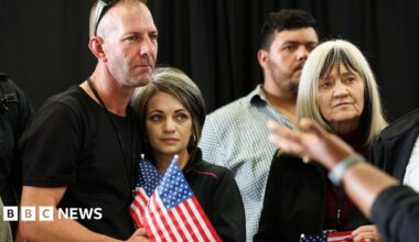 A man wearing sunglasses on his head with his arm around a woman. They are standing on the left and each holding a mini American flag. To their right, is an older woman. Behind her is a man. The background appears to be a black curtain.