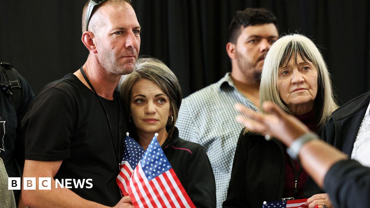A man wearing sunglasses on his head with his arm around a woman. They are standing on the left and each holding a mini American flag. To their right, is an older woman. Behind her is a man. The background appears to be a black curtain.
