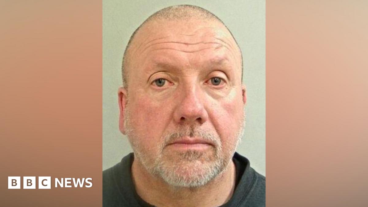 A mugshot of a middle aged man. He has a shaved head and a short grey beard. He is wearing a black T-shirt and  black sweater jumper.