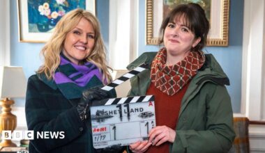 A woman with blonde hair and a woman with brown hair hold a clapper board between them which reads "SHETLAND". Ashley Jenson, the blonde woman, wears a dark green and purple scarf and a dark coat. Alison O'Donnell wears a patterned red scarf, a red jumper and green waterproof jacket. They are inside a formal looking room with baby blue wall panels and white outlines, with pictures hanging on the wall.