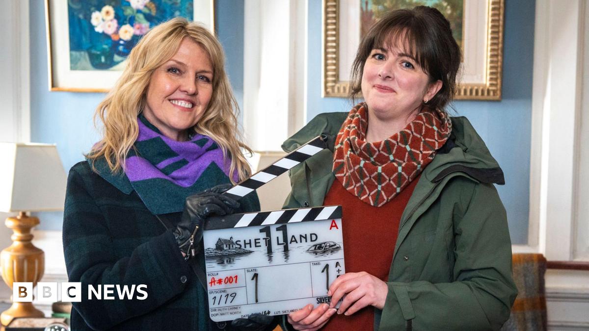 A woman with blonde hair and a woman with brown hair hold a clapper board between them which reads "SHETLAND". Ashley Jenson, the blonde woman, wears a dark green and purple scarf and a dark coat. Alison O'Donnell wears a patterned red scarf, a red jumper and green waterproof jacket. They are inside a formal looking room with baby blue wall panels and white outlines, with pictures hanging on the wall.