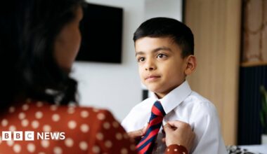 A woman is putting on her young boy's red and blue stripped tie for school. She is wearing an orange and white polka dot blouse and has brown hair her back is to the camera is blurred. The boy has brown hair and is wearing a white shirt