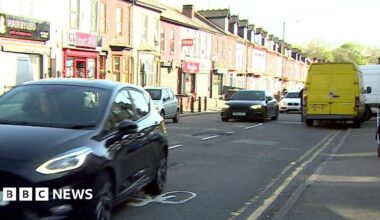 A street scene showing a row of terraced houses, several of which have shops built into the bottom floor. There are cars driving down the road and some parked on the pavement