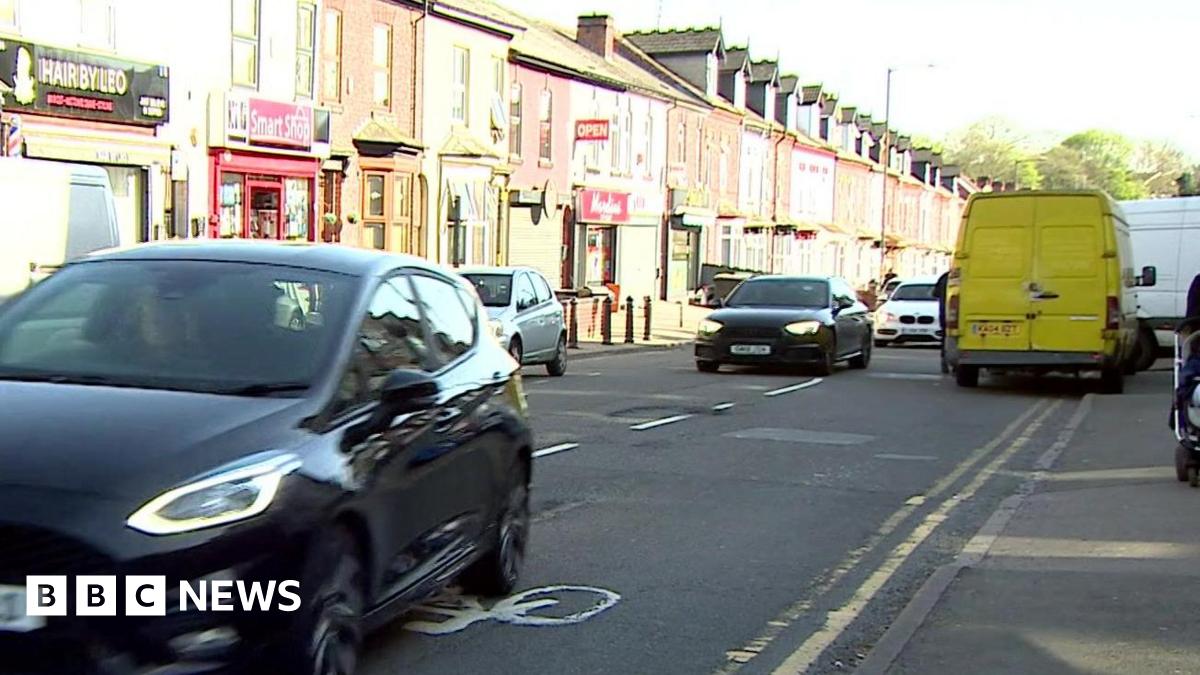 A street scene showing a row of terraced houses, several of which have shops built into the bottom floor. There are cars driving down the road and some parked on the pavement