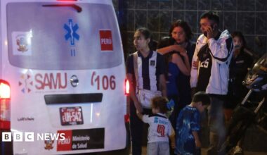 Crowds of people gathering near an ambulance at one of the entrances of the Alejandro Villanueva Stadium in Lima, Peru. It's night time and some of the fans are hugging, others looked shocked.