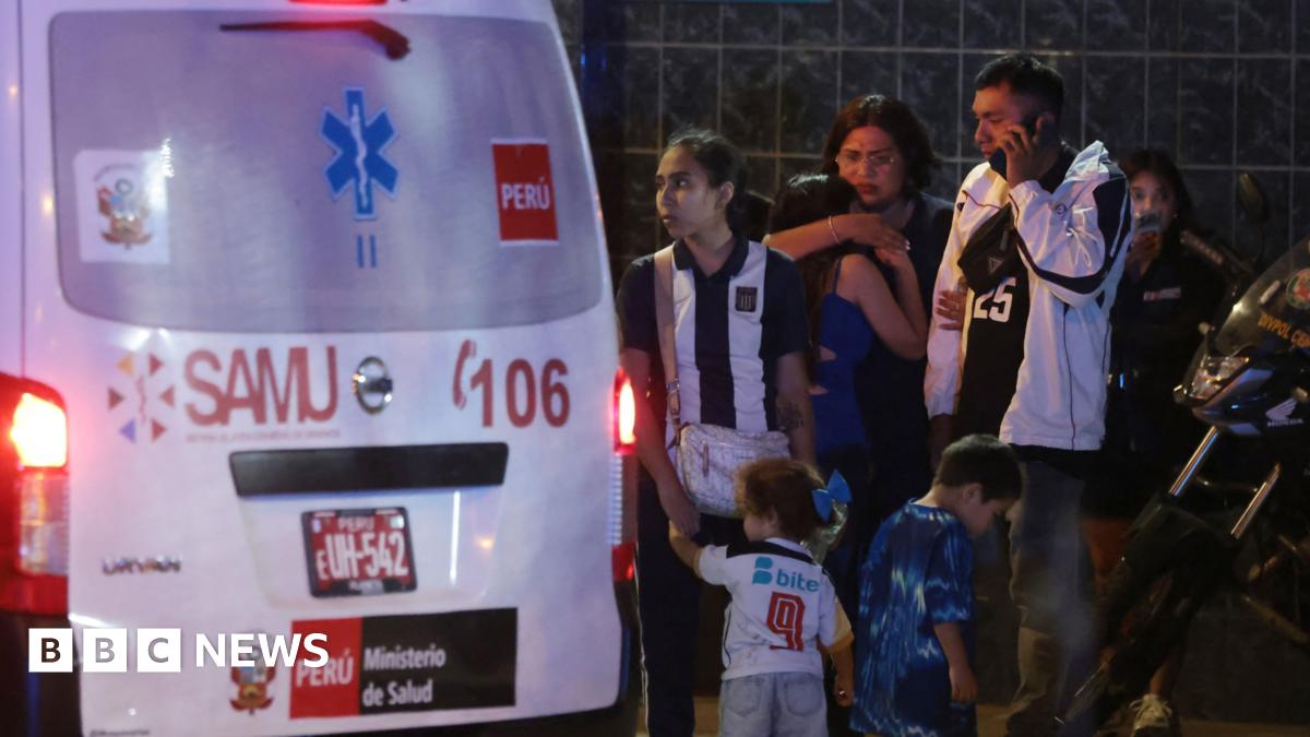 Crowds of people gathering near an ambulance at one of the entrances of the Alejandro Villanueva Stadium in Lima, Peru. It's night time and some of the fans are hugging, others looked shocked.