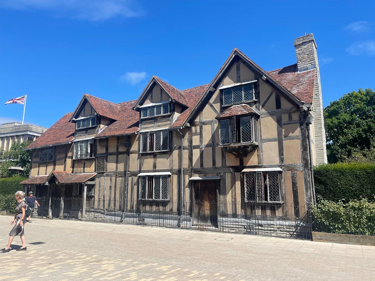 A historic half-timbered house with steep gabled roofs and small windows, situated in a sunny, open area with a couple walking past