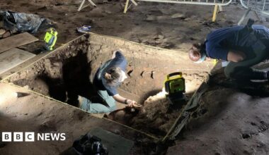 Archaeologists at a site are digging in a rectangular hole in the ground surrounded by metal fences. They are wearing head torches