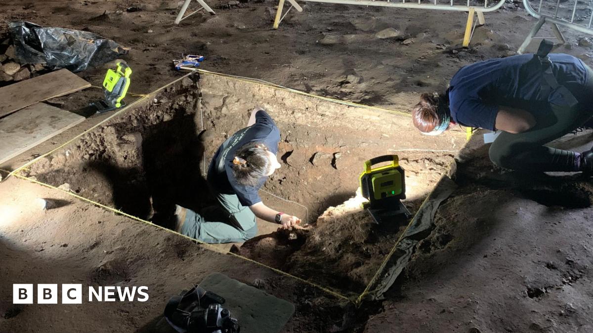 Archaeologists at a site are digging in a rectangular hole in the ground surrounded by metal fences. They are wearing head torches