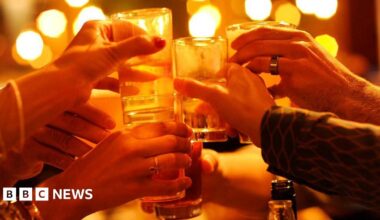 A group of people toast their drinks together in front of some lights. Their hands can be seen holding glasses.