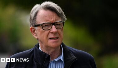 Lord Mandelson has short grey hair swept to one side and is wearing black-rimmed glasses. He has a dark blue jacket on and white and blue striped shirt underneath