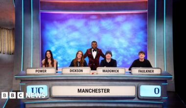 Four students sat at a table. There are name tags on the table (left to right POWER, DICKINSON, MADGWICK, FAULKNER). There is the word Manchester and UC on the front of the table. Behind the four sat down students is the presenter standing up. He is wearing a dark red suit.