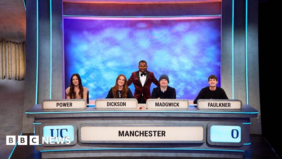 Four students sat at a table. There are name tags on the table (left to right POWER, DICKINSON, MADGWICK, FAULKNER). There is the word Manchester and UC on the front of the table. Behind the four sat down students is the presenter standing up. He is wearing a dark red suit.