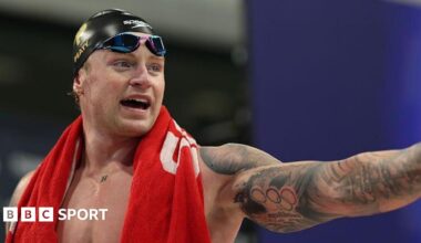 Adam Peaty in the swimming pool with a red swimming cap on. He is leaning on a floating lane divider