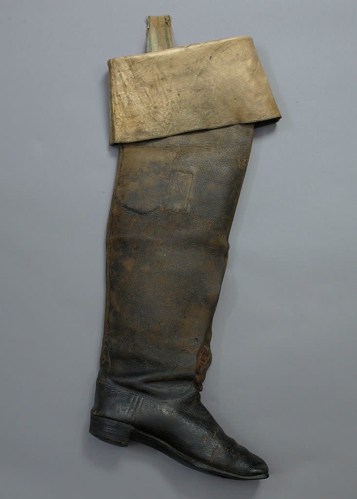 A well-worn, tall leather boot with a folded top and a strap, possibly belonging to an adult, displayed vertically