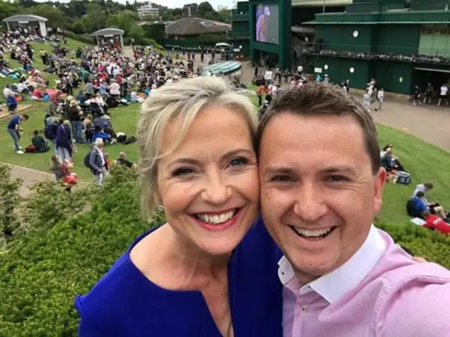 Carol and Matt smiling together outdoors at Wimbledon with crowd seated on hill behind them on a bright summer day