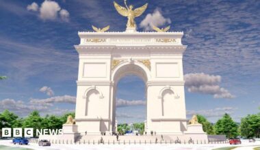 David Robinson is photographed indoors against a pale wall with a subtle floral pattern. He is shown from the shoulders up, wearing a light shirt and a dark jacket. The lighting is soft and even, and the background is uncluttered.