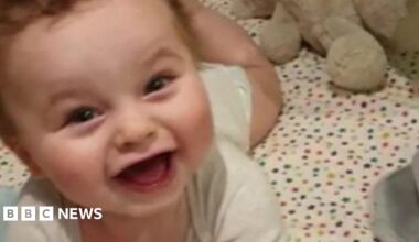 Baby Preston Davey smiles at the camera dressed in a white vest and appears to be lying in a cot with star bedsheet cover