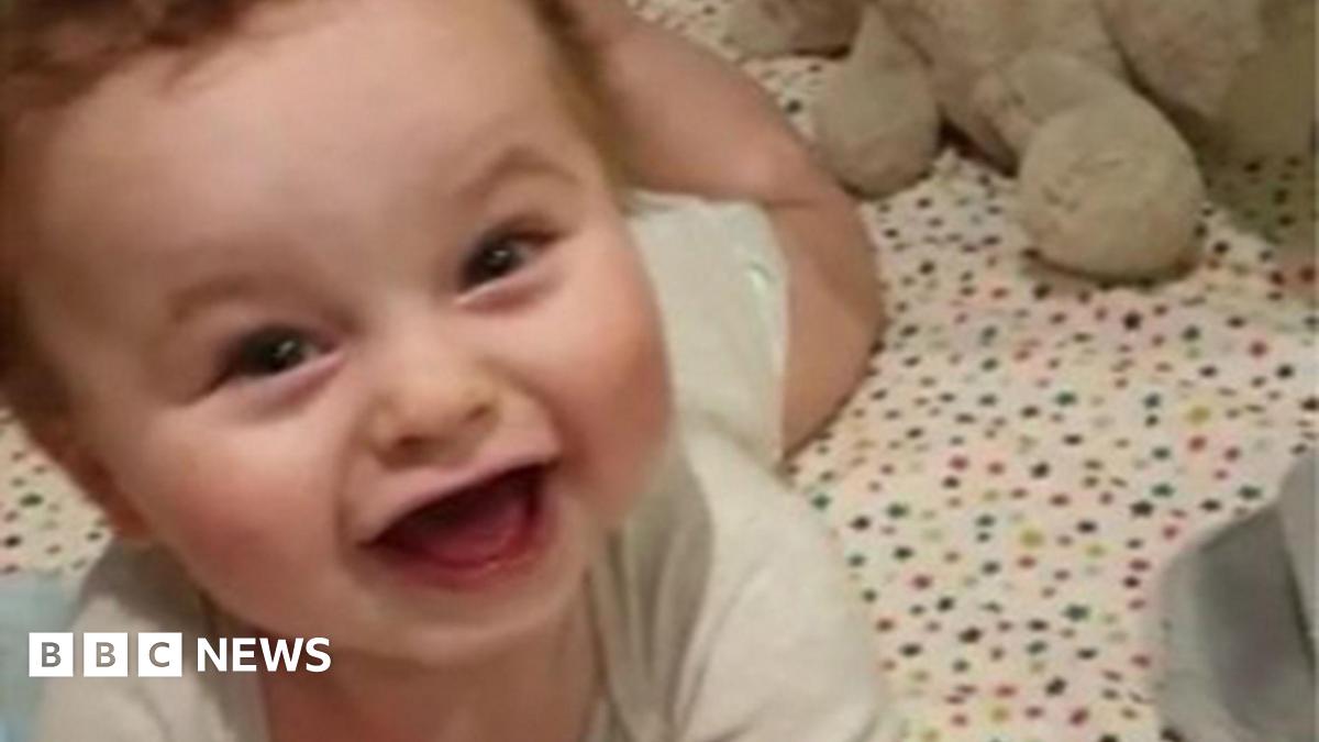 Baby Preston Davey smiles at the camera dressed in a white vest and appears to be lying in a cot with star bedsheet cover
