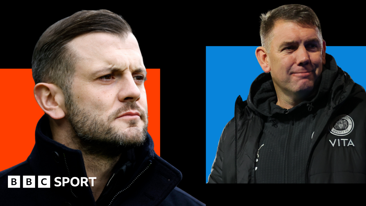 Jack Wilshere, wearing a black coat with a high collar, looks on with a serious face (left). Dave Challinor, wearing a black jacket with a Stockport county badge on, smiles (right).