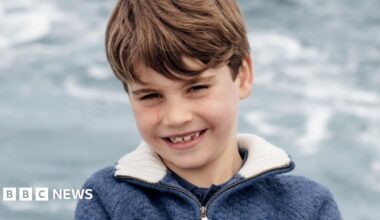 A grinning Louis is shown leaning with his arms folded against a metal barrier, with the sea as the backdrop.
