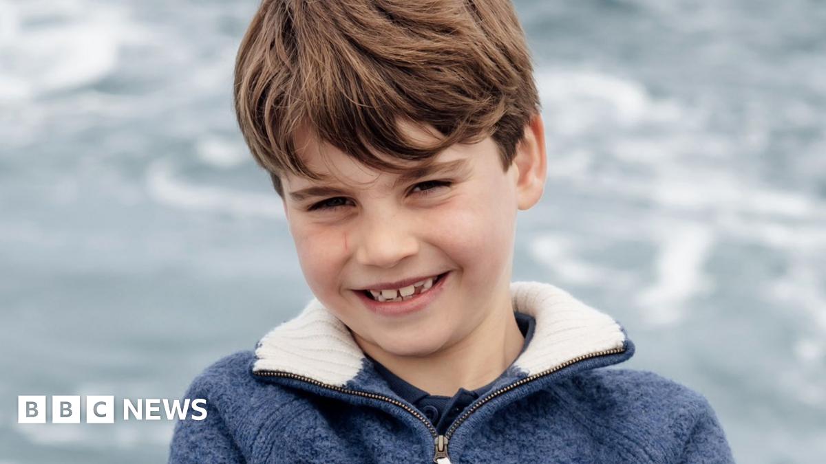 A grinning Louis is shown leaning with his arms folded against a metal barrier, with the sea as the backdrop.