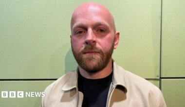 Paul Cheema is a man in his 50s wearing glasses with grey and black hair. He is standing in his petrol station in front of the tills and he is holding illegal tobacco products including a packet of 20 cigarettes and a bag of tobacco.