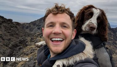 David Parrish a man with curly hair and a beard smiles into the camera with his dog leaning on his shoulder