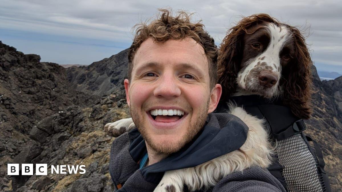 David Parrish a man with curly hair and a beard smiles into the camera with his dog leaning on his shoulder