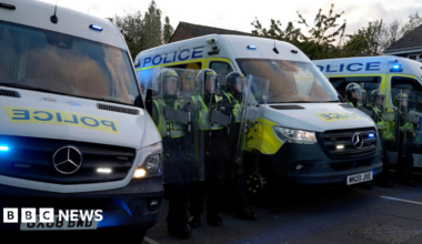 Police officers with riot shields stand in a line next to police vans.