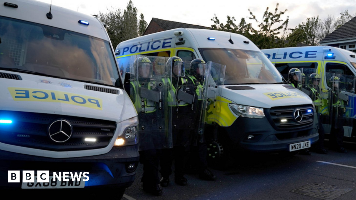 Police officers with riot shields stand in a line next to police vans.