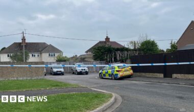 The police cordon on Butler's Field in Lechlade. There is police tape across the road, with a police car parked to one side. There is a car park, with two vehicles parked in it and residential properties in the distance.
