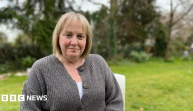Linda Franklin sits on a chair in a garden surrounded by trees. She has a blond bob and brown eyes and wears a grey cardigan, with a white top underneath.