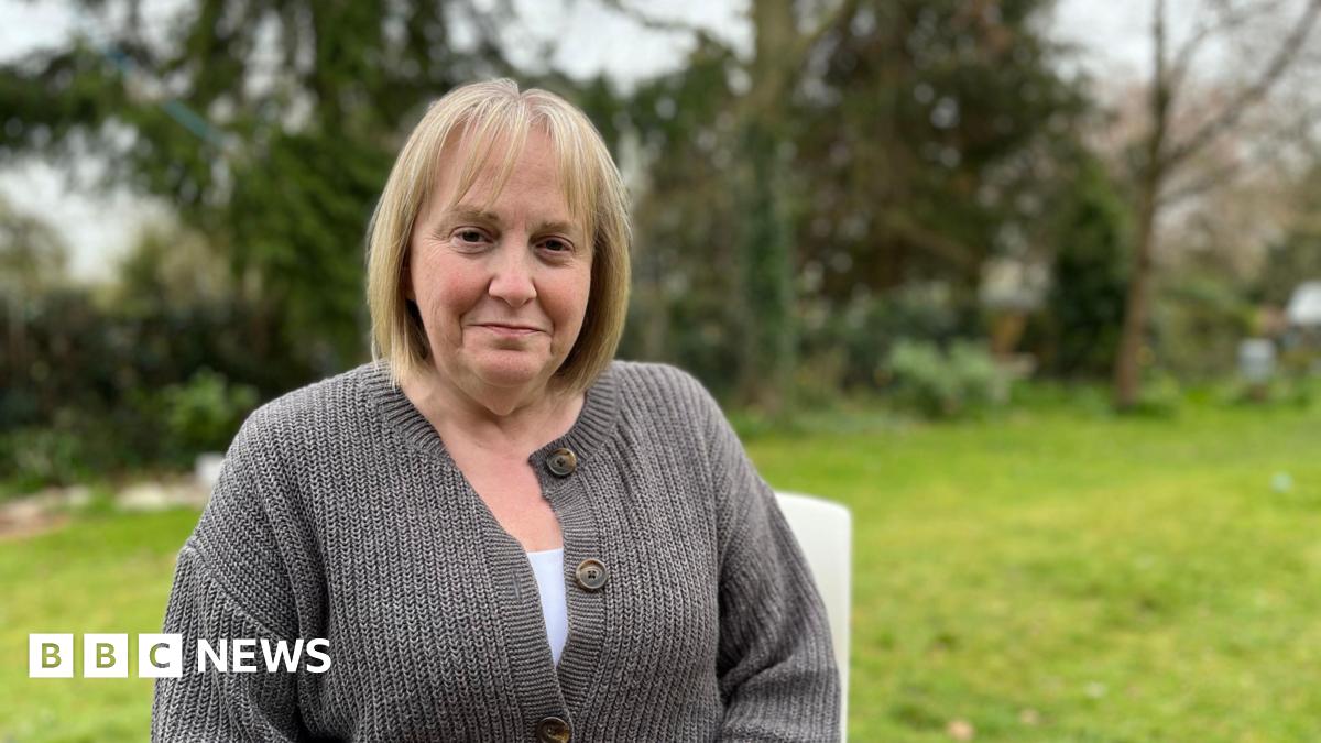 Linda Franklin sits on a chair in a garden surrounded by trees. She has a blond bob and brown eyes and wears a grey cardigan, with a white top underneath.