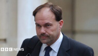 A man with brown hair and a moustache who is wearing a black suit and tie with a white shirt