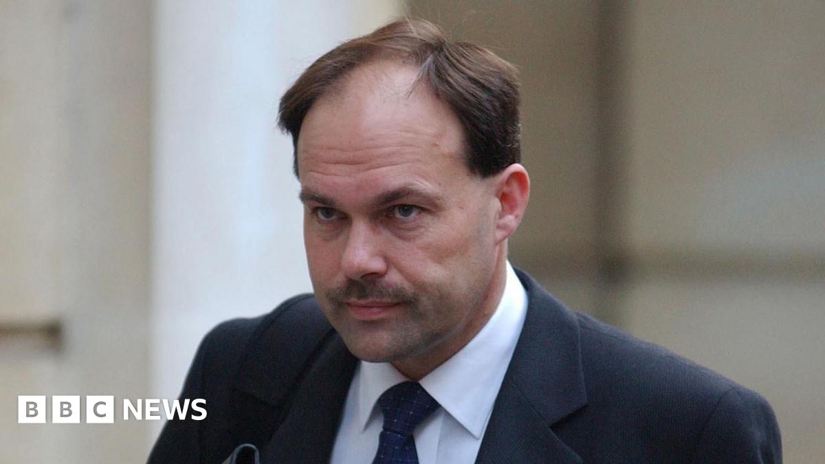 A man with brown hair and a moustache who is wearing a black suit and tie with a white shirt