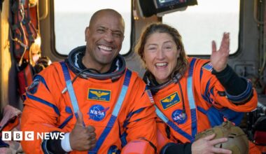 Four astronauts in dark blue T‑shirts float close together inside a bright, cluttered spacecraft. Their faces are blurred, but all give thumbs‑up to the camera. Their shirts carry a matching mission patch and small US flag on the sleeve. Behind them, American and Canadian flags hang above a red‑and‑blue “America 250” logo. Around them are white padded walls, orange pipes, cables and tightly netted blue and orange cargo bags, emphasising the cramped zero‑gravity cabin.