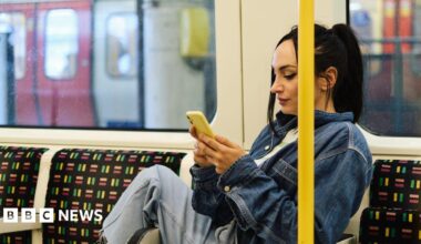 A woman wearing jeans and a denim jacket scrolls on her smartphone whilst on the Tube in London.